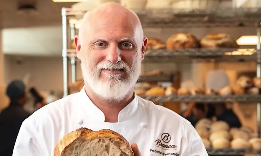 Federico Fernandez in a white chef's jacket holding loaves of sourdough in front of bread racks