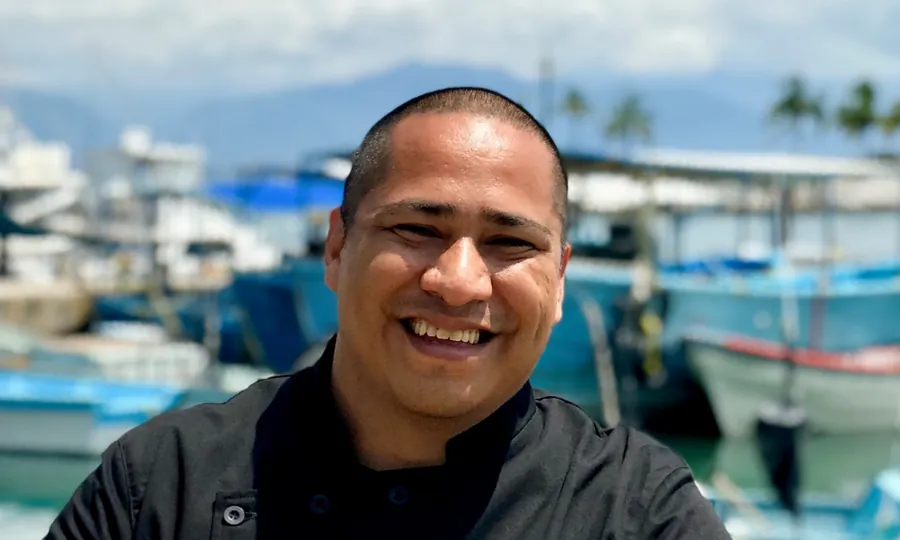Héctor Leyva smiling in a black chef's jacket on a dock, with fishing boats behind him