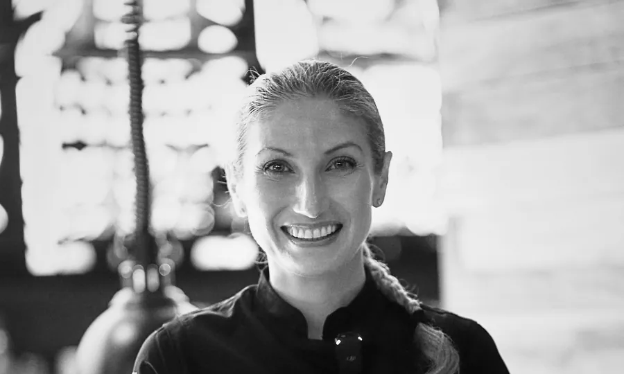 Black-and-white portrait of Leslie Durso smiling, in a dark chef's jacket, hair tied back