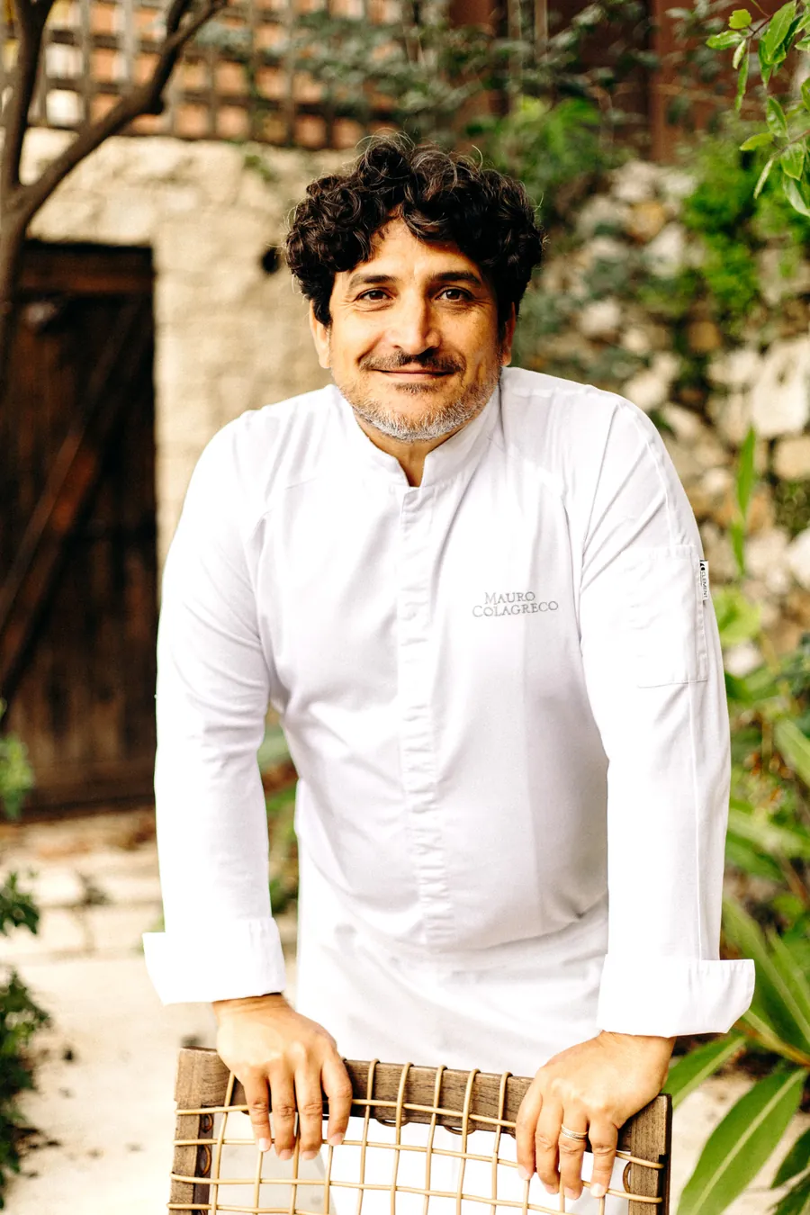 Mauro Colagreco in a white chef's jacket, leaning on the back of a wicker chair beside an old wooden door and a stone wall