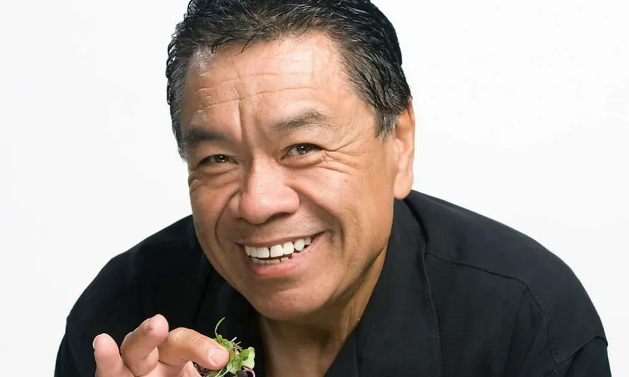 Sam Choy laughing in a black shirt, holding up a small plate of food