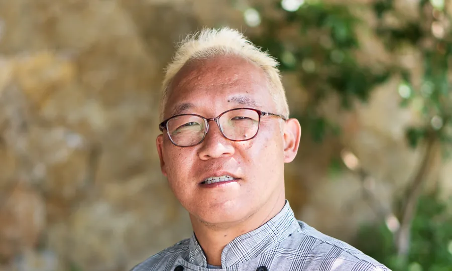 Yasuo Asai in glasses and a grey patterned jacket, smiling, with foliage behind