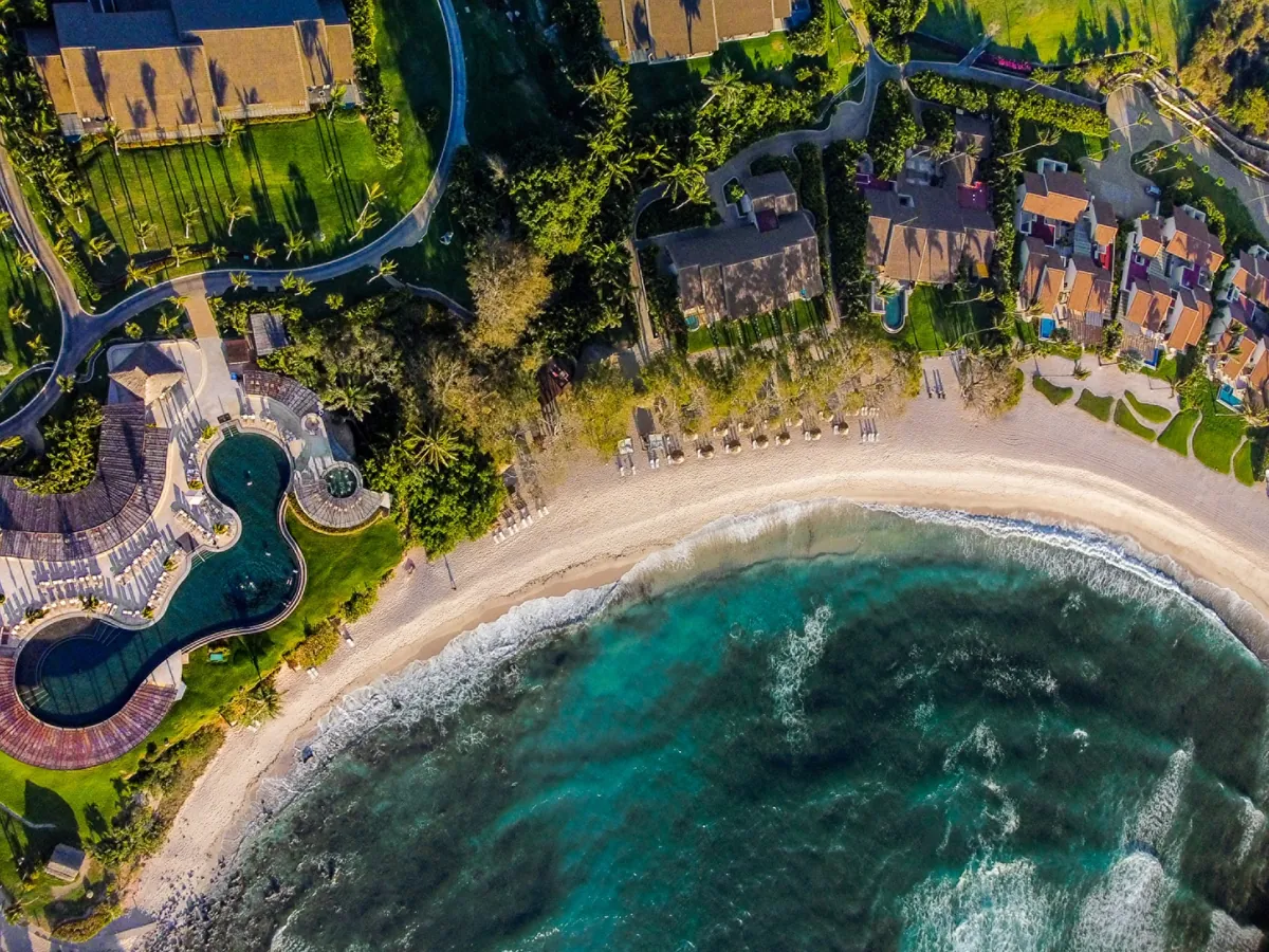Aerial view straight down on a curved sandy beach, turquoise water, free-form pools, palms and tiled roofs at Four Seasons Resort Punta Mita