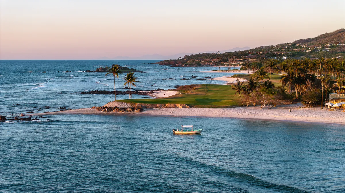 A golf fairway running along a curved sandy beach at sunset, palm trees on the shore and a small boat on the sea, Punta Mita
