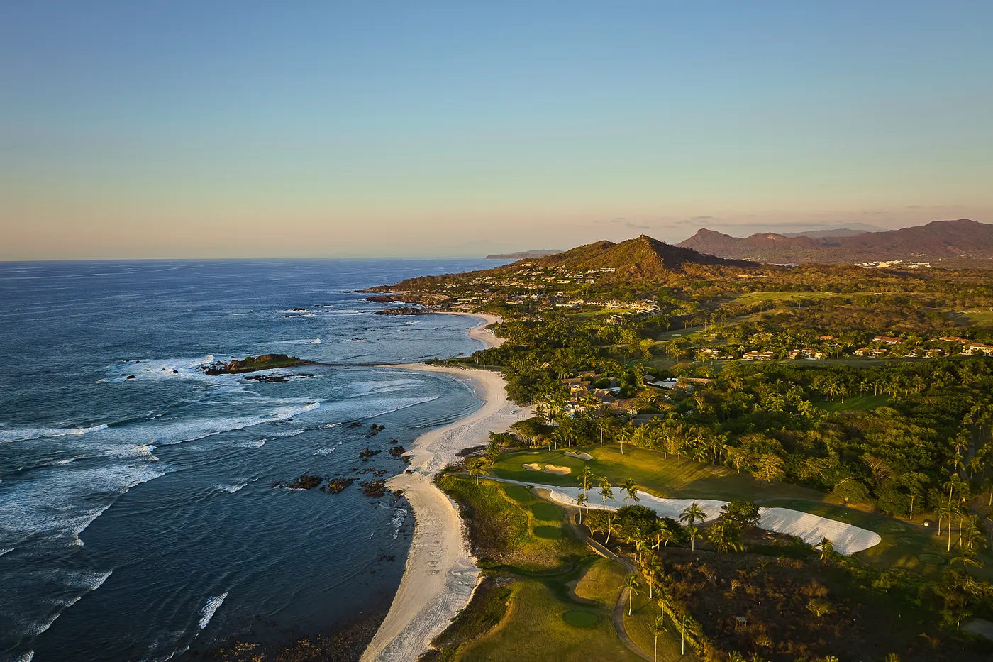 Aerial view at dusk of green fairways and bunkers along a curved beach, with the Tail of the Whale island green in the sea and hills behind, Punta Mita