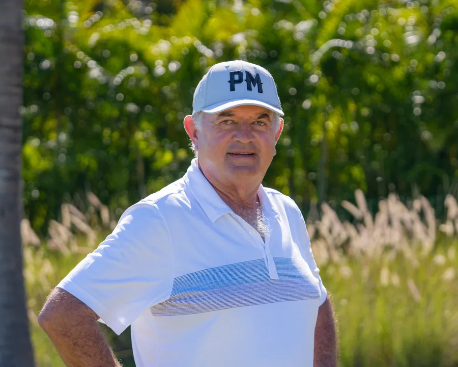 Rafael Alarcón in a white polo and a PM cap, standing among tall grasses and palms