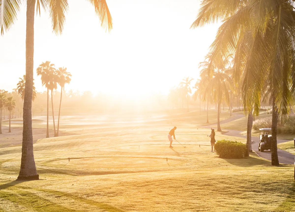 Two golfers on a tee between tall palm trees at sunrise, backlit, with a golf cart parked at the right, Punta Mita