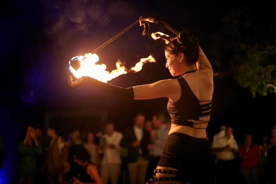 A fire dancer swinging flaming poi at night in front of a crowd of guests