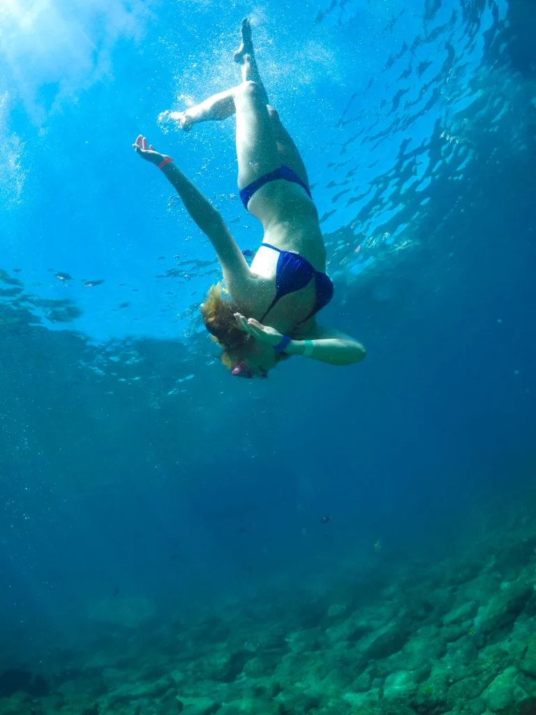 A snorkeller dives head-down through clear blue water towards a pale sandy seabed.