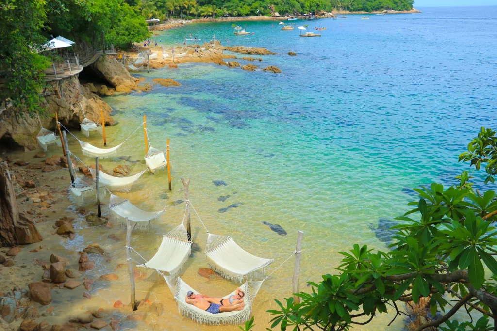 White hammocks strung on posts above turquoise shallows at the edge of a rocky sand beach.