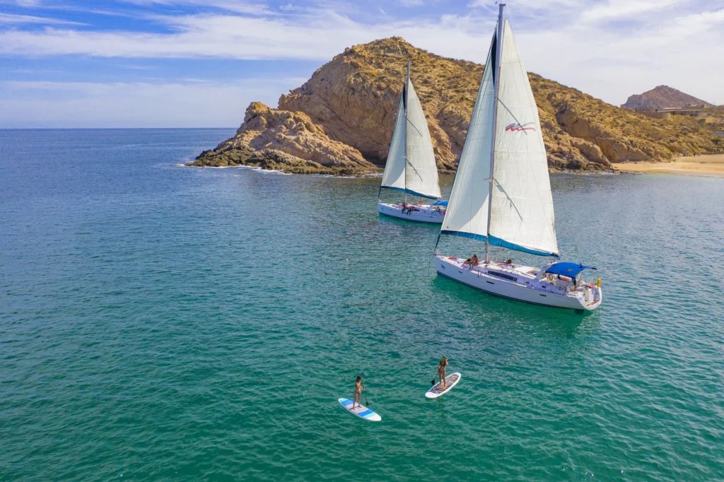 Two sailing boats lie off a rocky headland with paddleboarders on green water.
