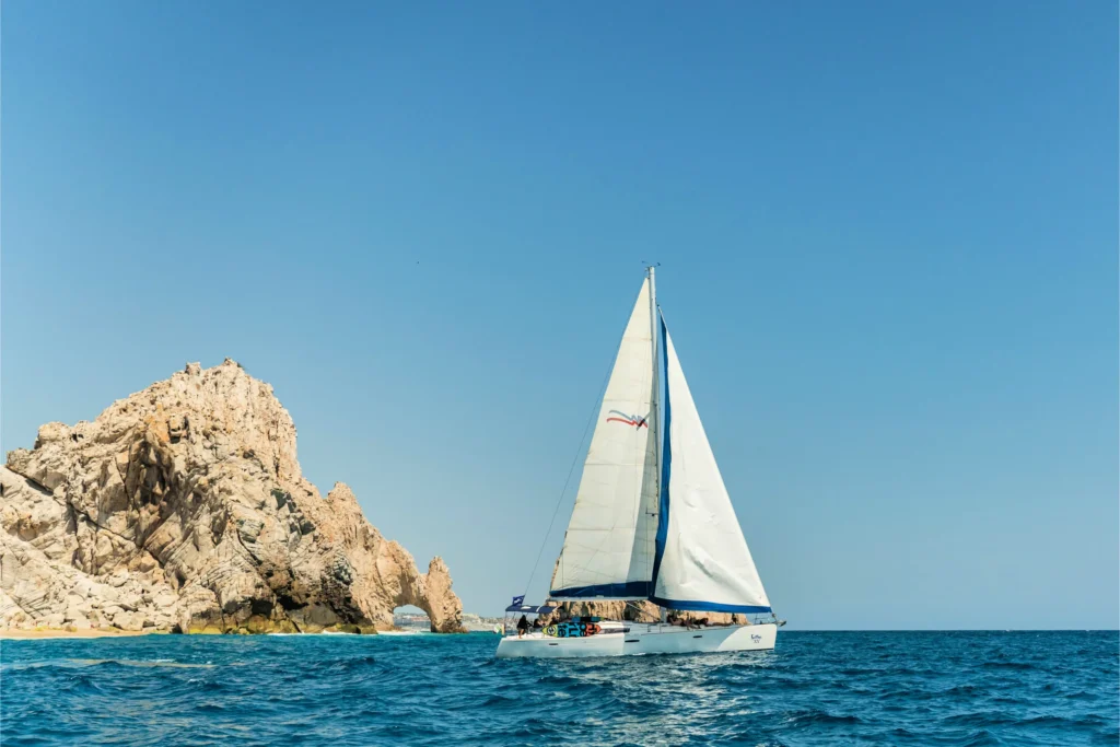 A sailing boat under white sail passes the Arch at Land’s End on deep blue water.