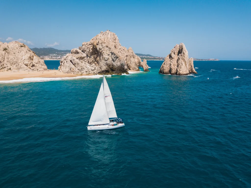 A sailing boat crosses in front of the pale sand of Lover’s Beach and the rocks.