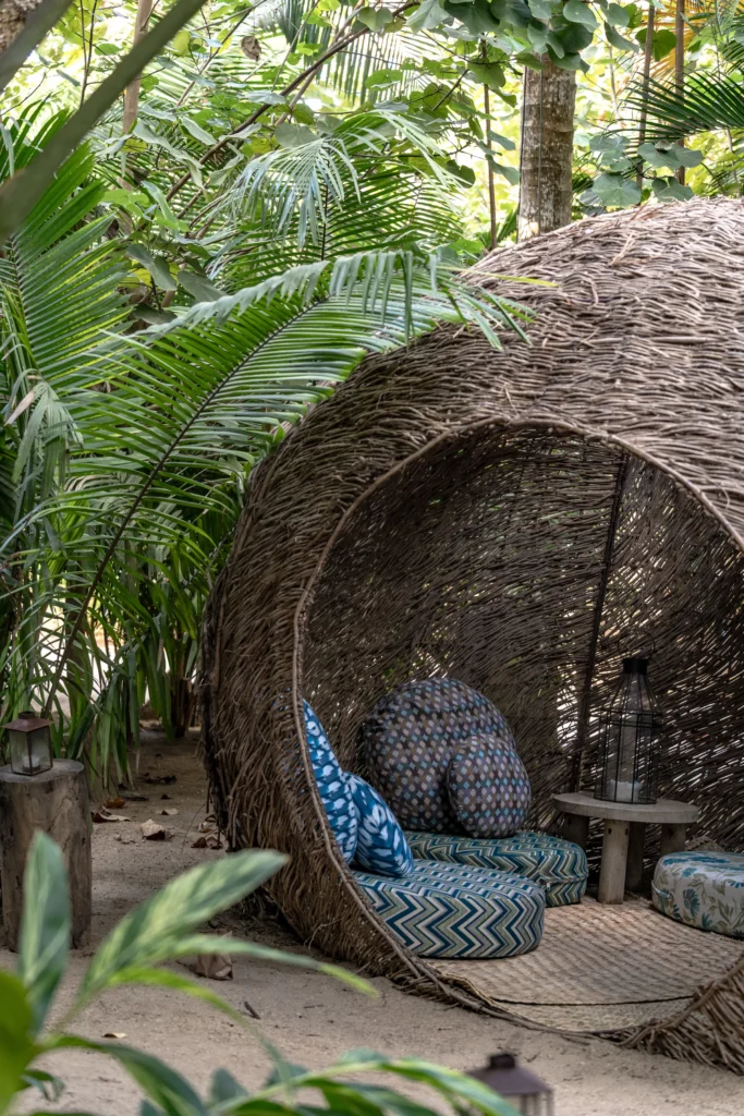 A large woven nest shelter holding patterned cushions sits on a deck among tall palms.