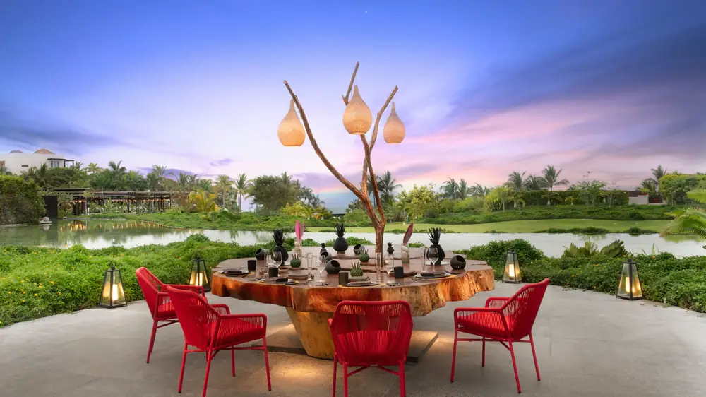 Mesa 1 at W Punta de Mita: a round table with red chairs under a sculpted tree hung with lamps, by the lake at dusk