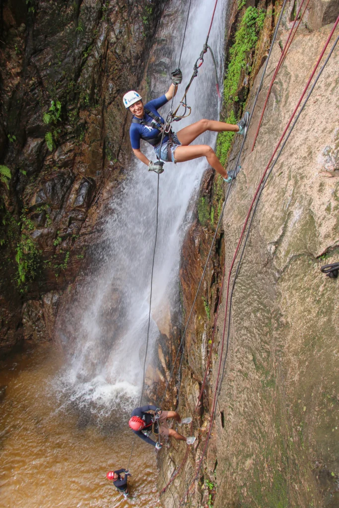 A person on a rope descends a dark rock face beside a long white waterfall in the jungle.