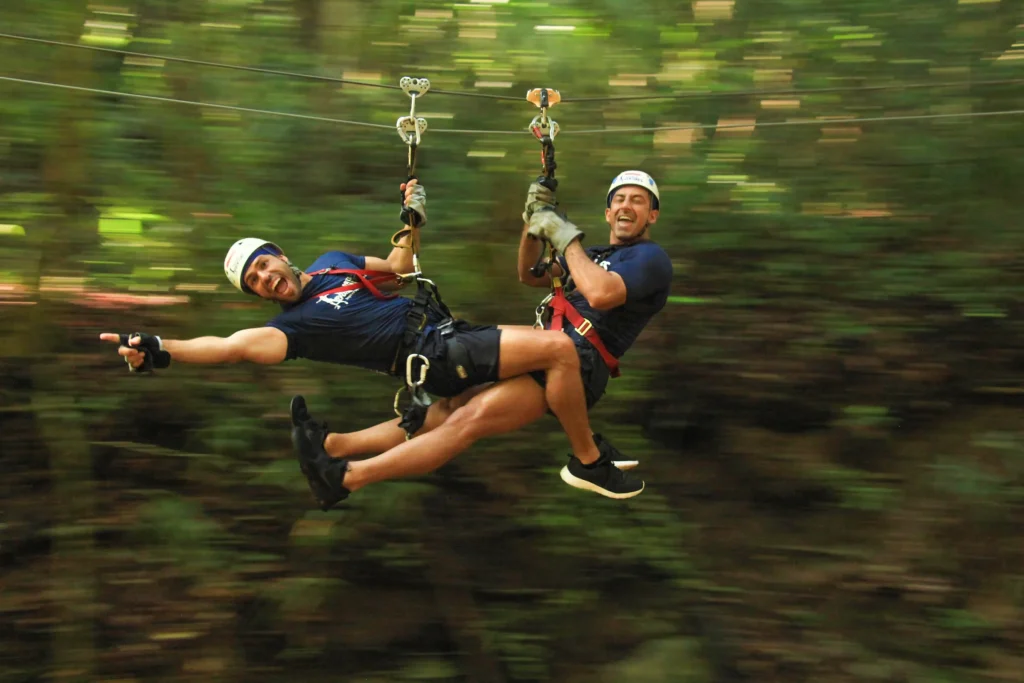 Two riders rush along a zipline, the green jungle behind them blurred by the speed.