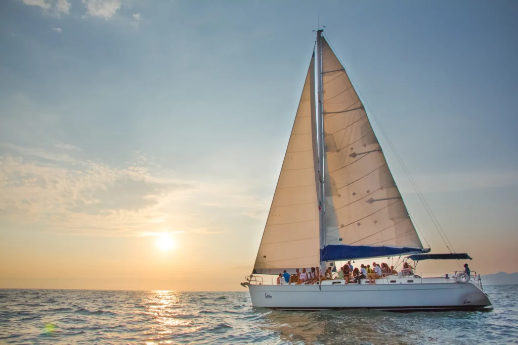 A catamaran under full white sail heads towards a low sun on a bright silver sea.