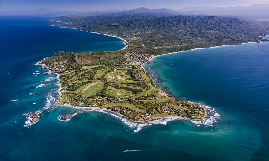 High aerial view of the Punta Mita peninsula: golf fairways across the point, white beaches on both sides and the Sierra behind