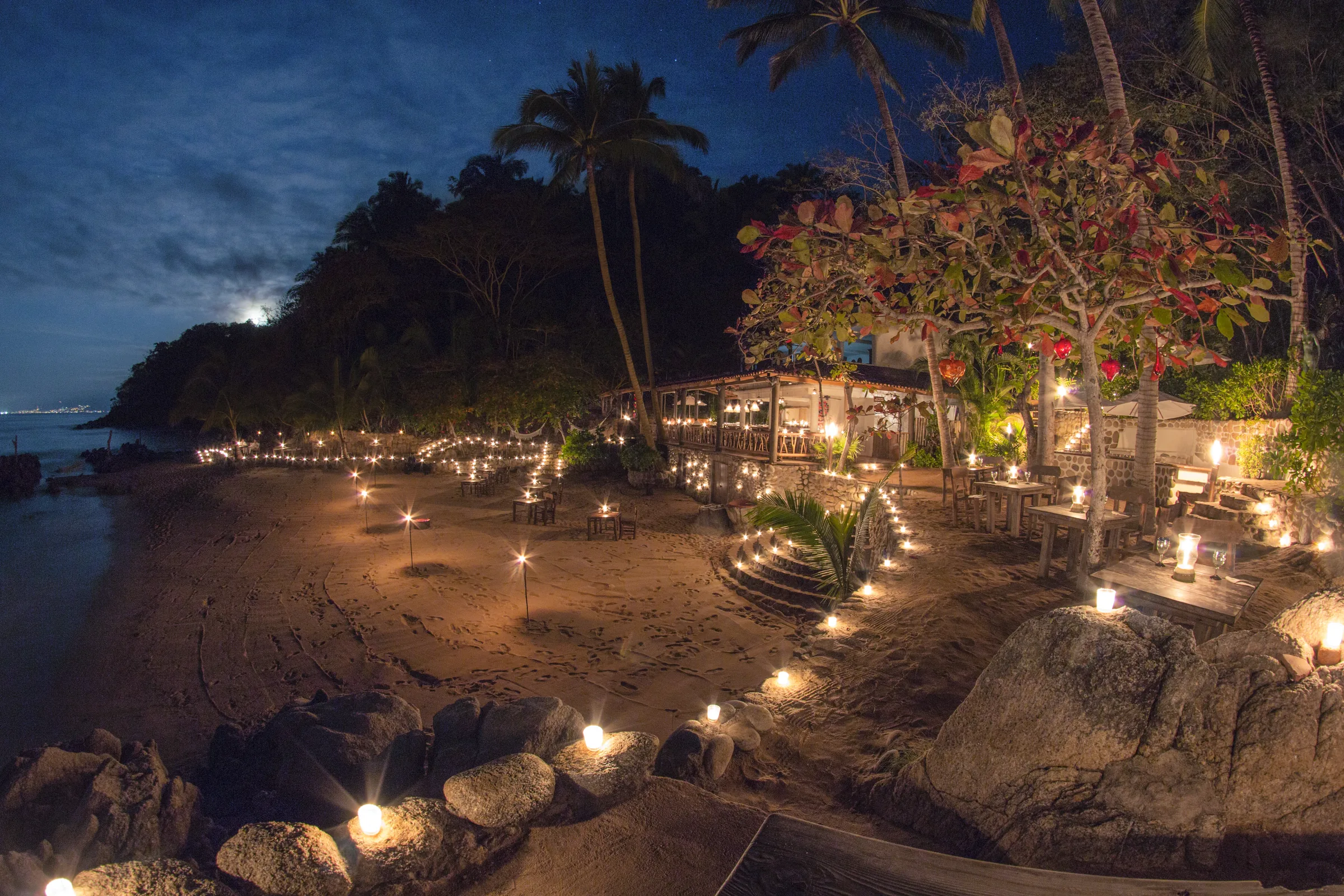 A curved beach lined with candles and lanterns under palms, lights reflecting on still water.