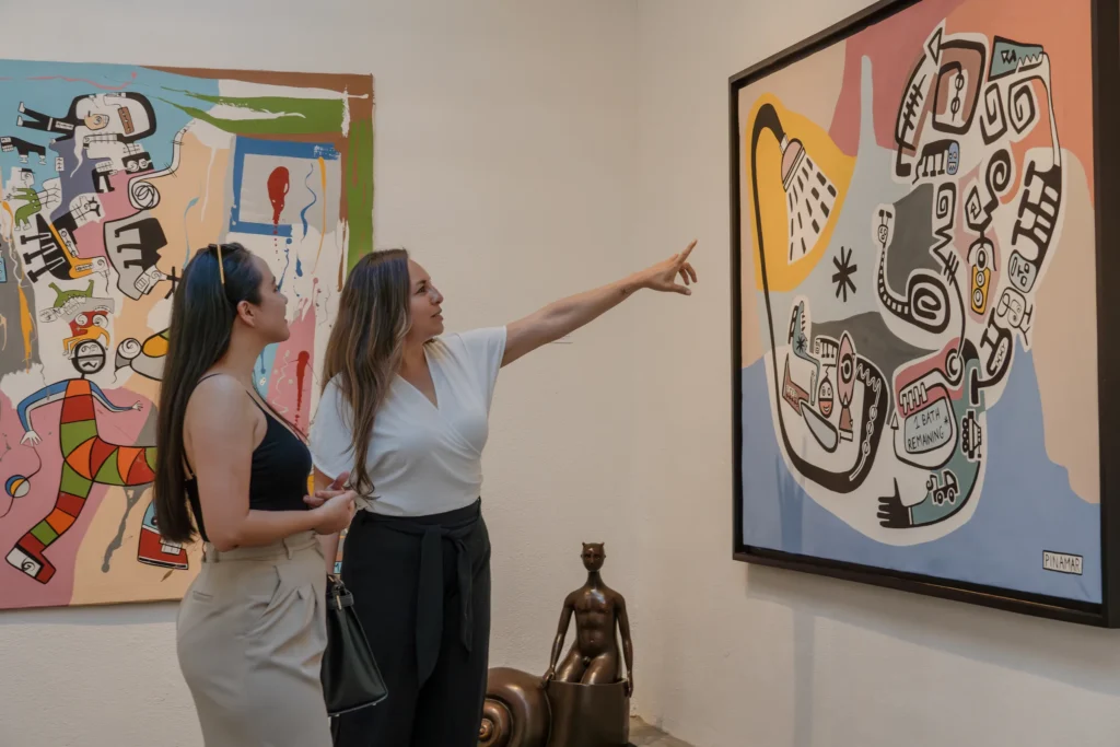 Two visitors stand in a gallery looking up at large bright paintings.