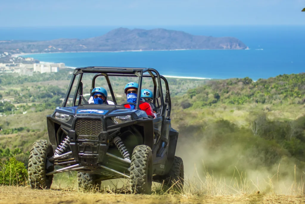 A buggy sits on a dirt track high above green scrub with the sea and a headland beyond.