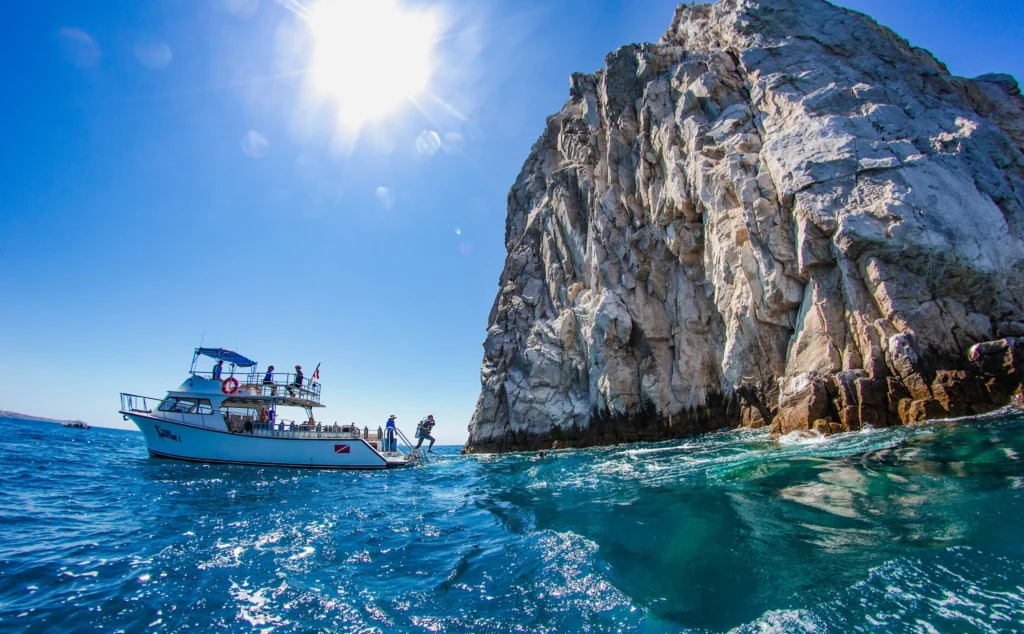 A dive boat sits on blue water below a tall pale granite rock.