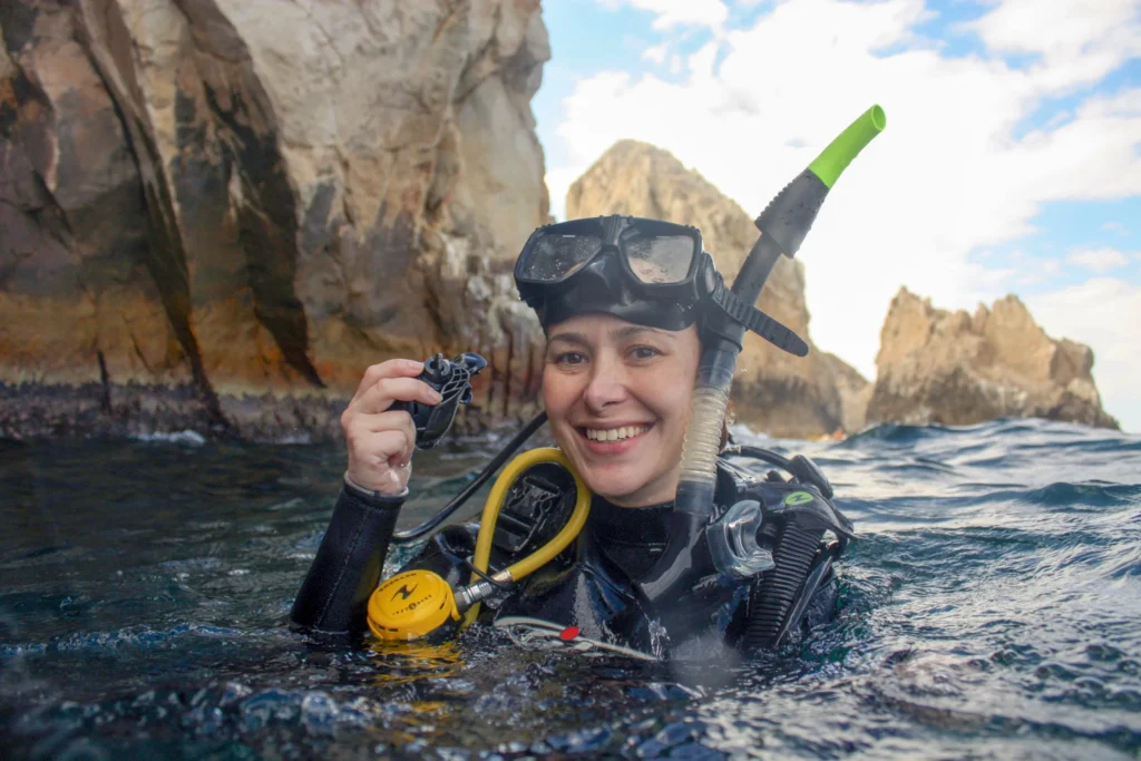 A diver at the surface holds a camera with the Land’s End rocks behind.