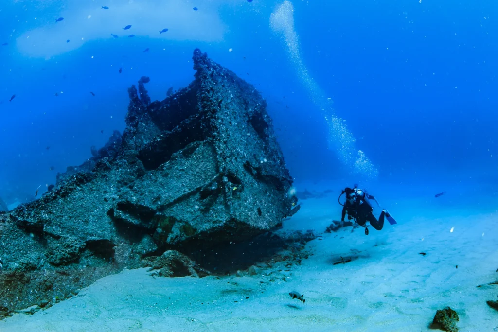 Two divers swim beside the dark shape of a wreck on the bottom.