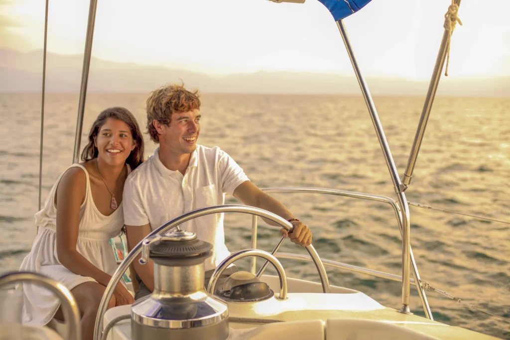 Two guests stand at the wheel of a sailing boat in warm evening light.
