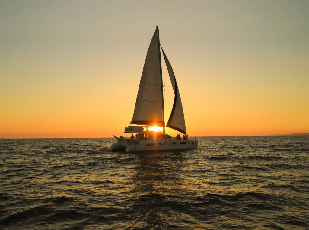 A sailing catamaran crosses the frame with the low sun burning through its sail.