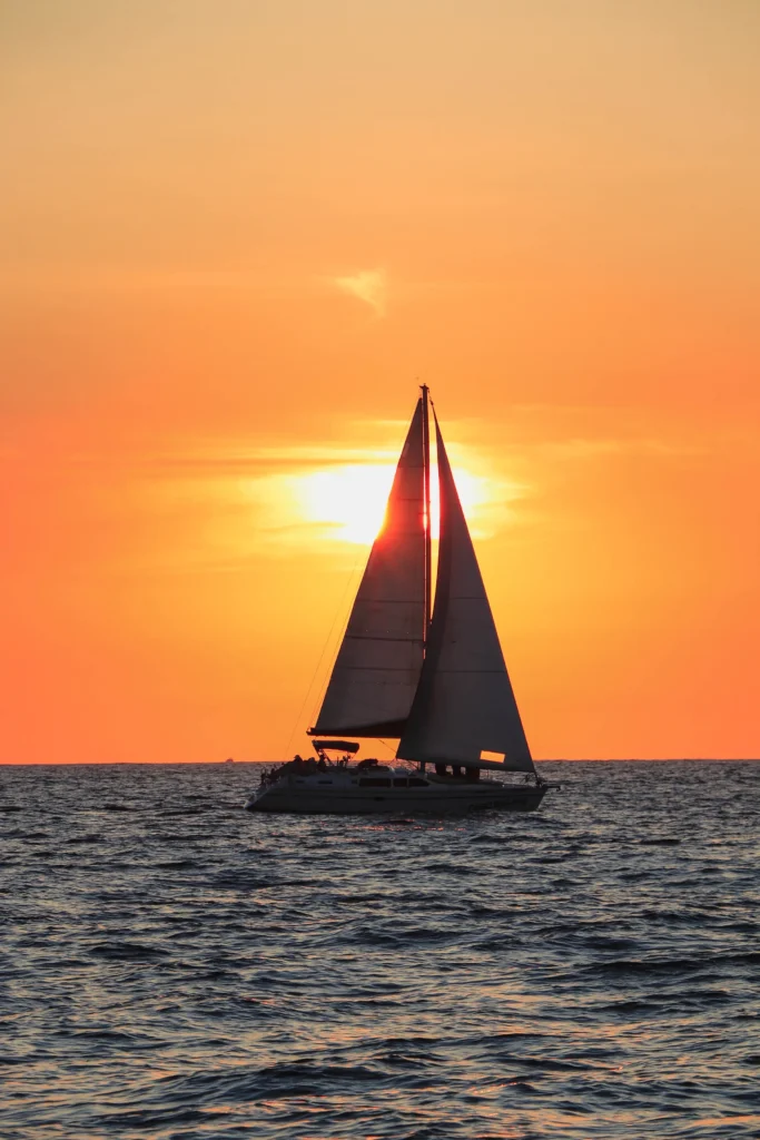 A sail is silhouetted against a large orange sun low on the horizon.