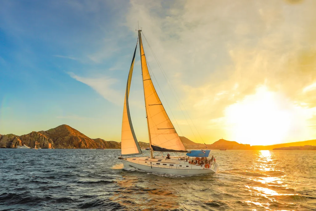 A sailing boat passes a headland with the setting sun glowing through its sail.