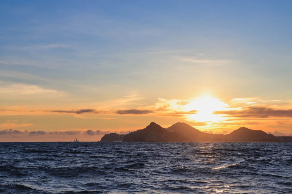 The sun sets behind a dark headland with calm water in the foreground.