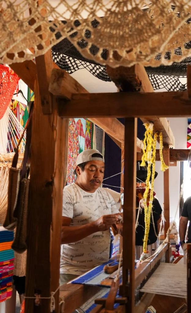 A weaver works a wooden floor loom in a workshop hung with woven goods.