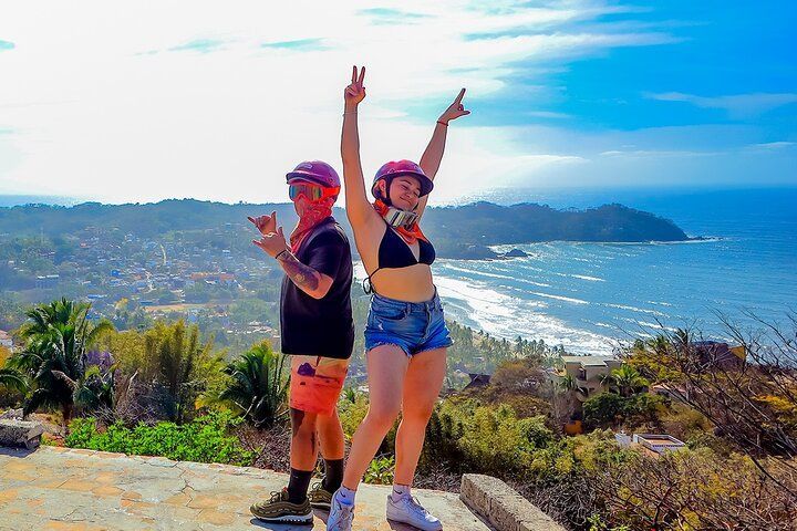 Two riders in helmets posing at a hilltop lookout above Sayulita's coastline