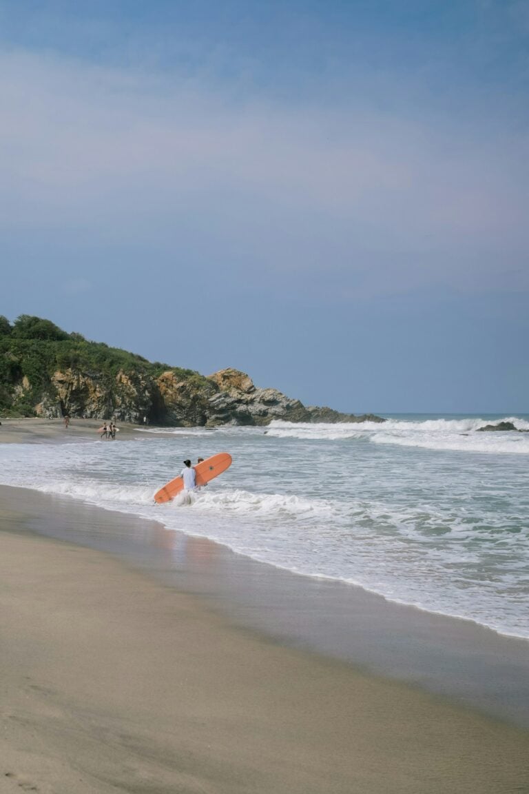 A surfer carries an orange board out of the waves on a wide Puerto Escondido beach with green headland behind