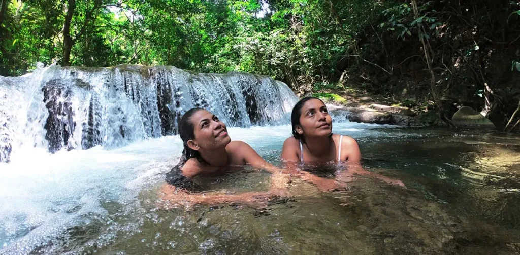 Two women bathing in a jungle pool beneath a wide, low waterfall