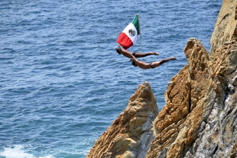 Two La Quebrada cliff divers in mid-air beside a Mexican flag, leaping from the rocks toward the Pacific