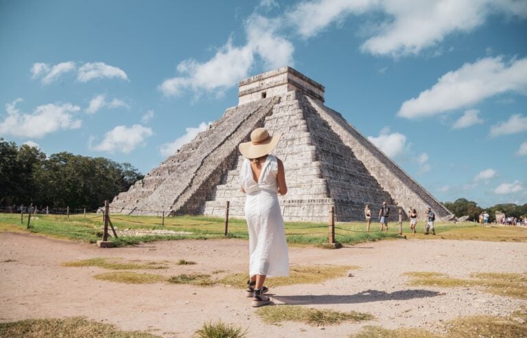 A woman in a white dress and sun hat looks up at the El Castillo pyramid at Chichen Itza