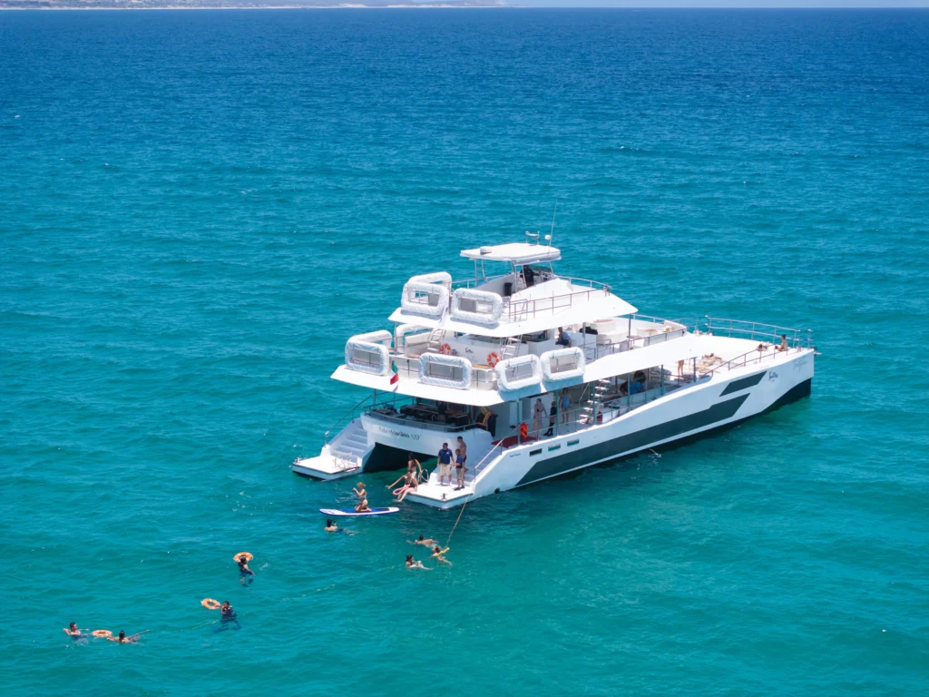 A catamaran lies at anchor over bright turquoise water with swimmers around it.