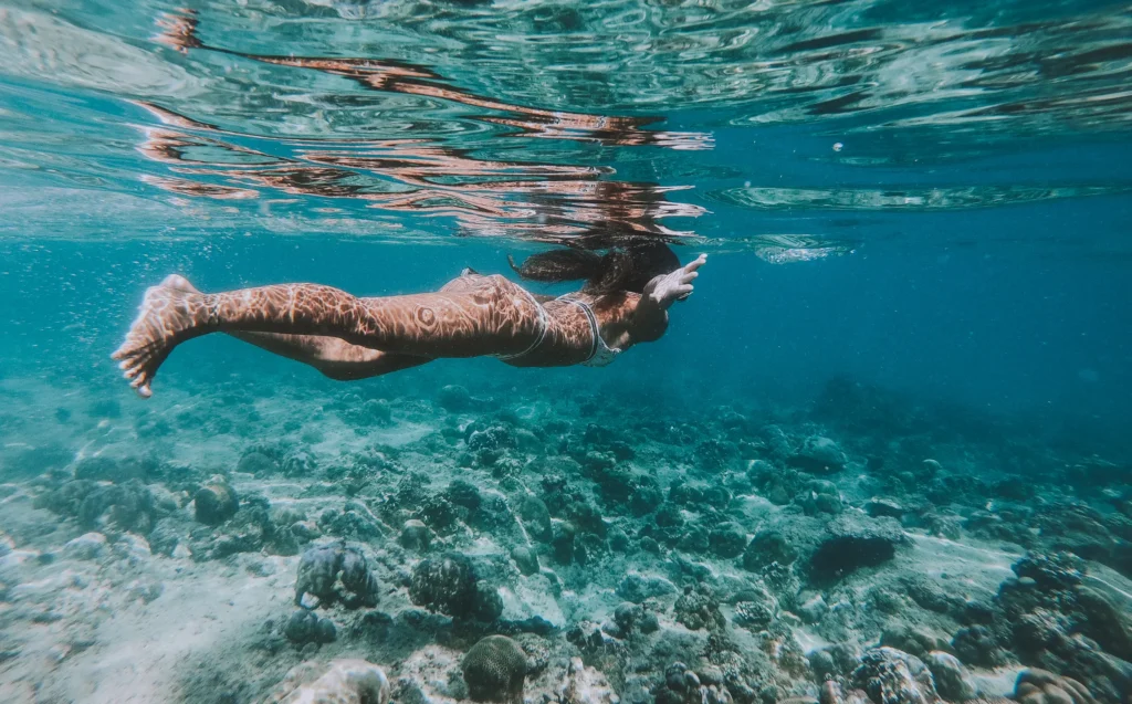 A snorkeller swims over pale rock and sand in bright shallow water.
