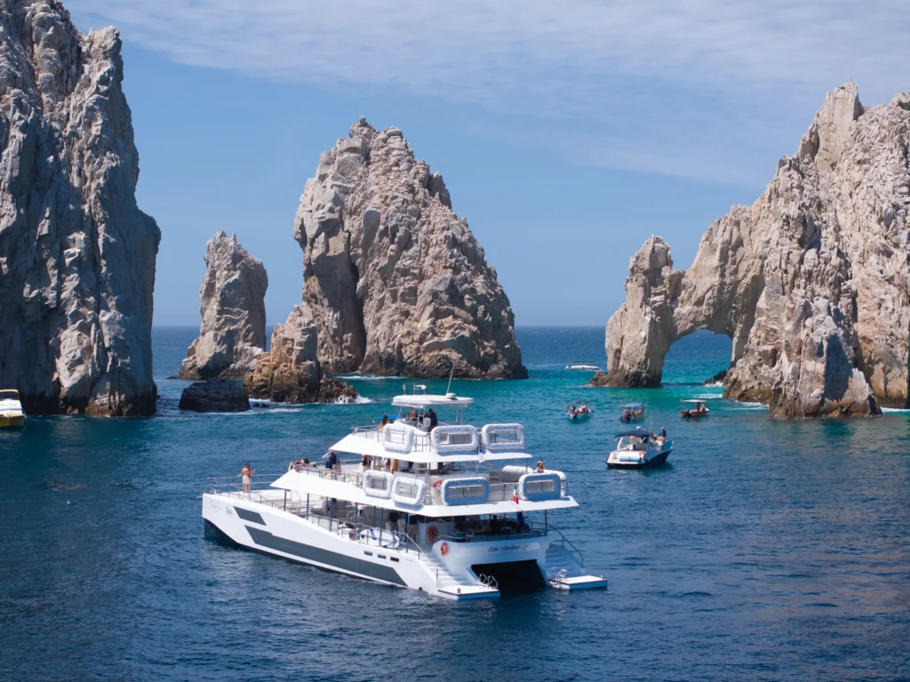 A large white catamaran sits on blue water in front of the Land’s End Arch.