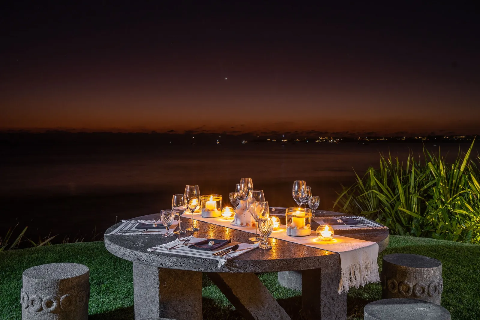 A round stone table set with candles and glasses on a lawn at night, the sea dark behind