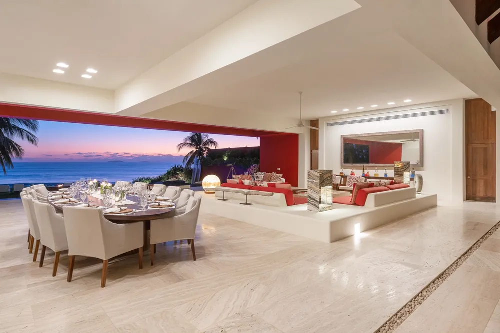 An open dining room with a long table and white chairs, open to a terrace and the sea at dusk