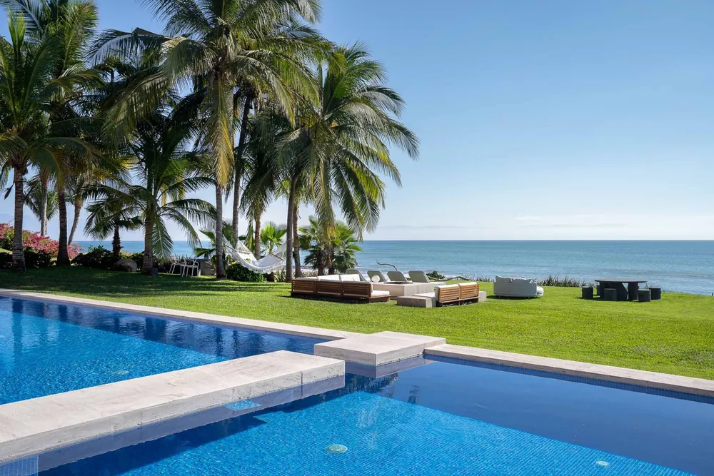 A long blue pool beside a lawn and palms, the sea beyond under a bright sky