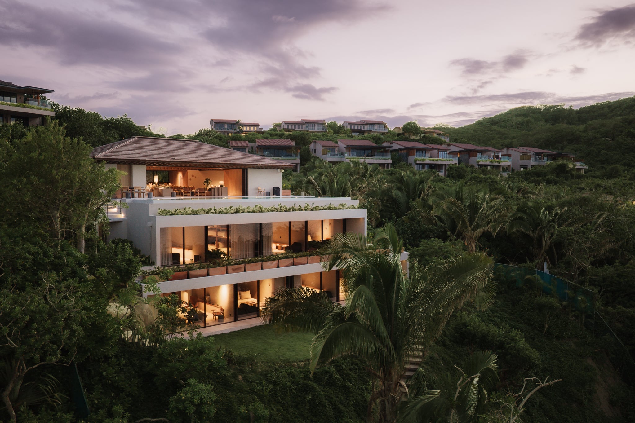 The three floors of Villa Taiyari lit at dusk above the lawn, the wooded hillside behind — Punta Mita Nayarit, Mexico