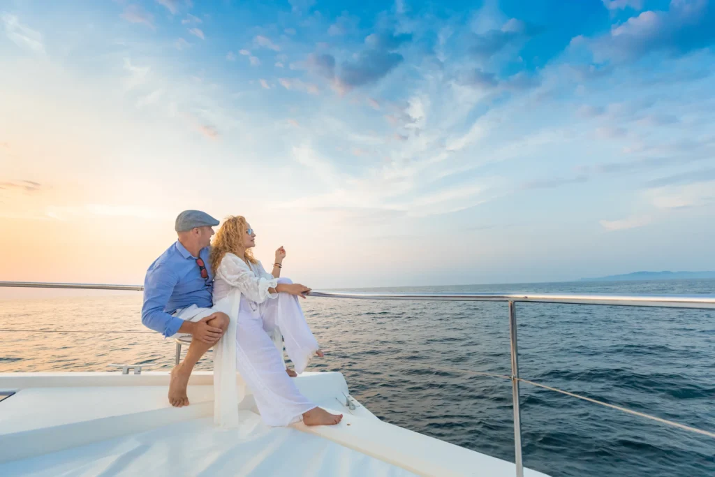 Two guests sit on the bow netting of a catamaran as the sun drops behind them.
