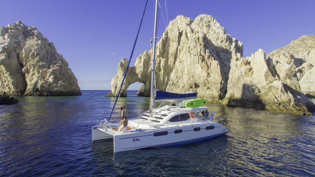 A sailing catamaran sits on blue water in front of the Land’s End rocks.