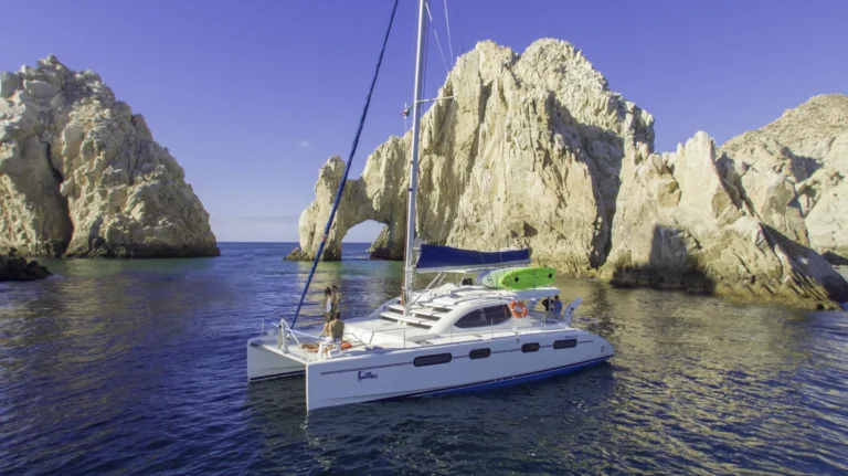 A sailing catamaran sits on blue water in front of the Land’s End rocks.